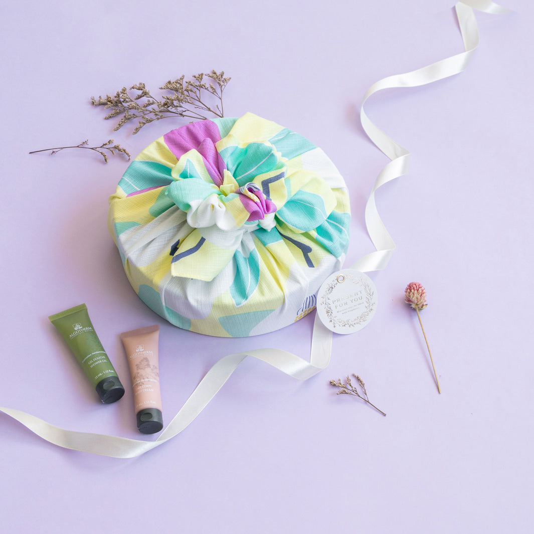 photo d'ambiance d'un emballage cadeau réalisé avec le furoshiki au motif de camélias dans les tons jaune clair, vert d'eau et rose pourpre adeline x musubi en 70cm par 70cm