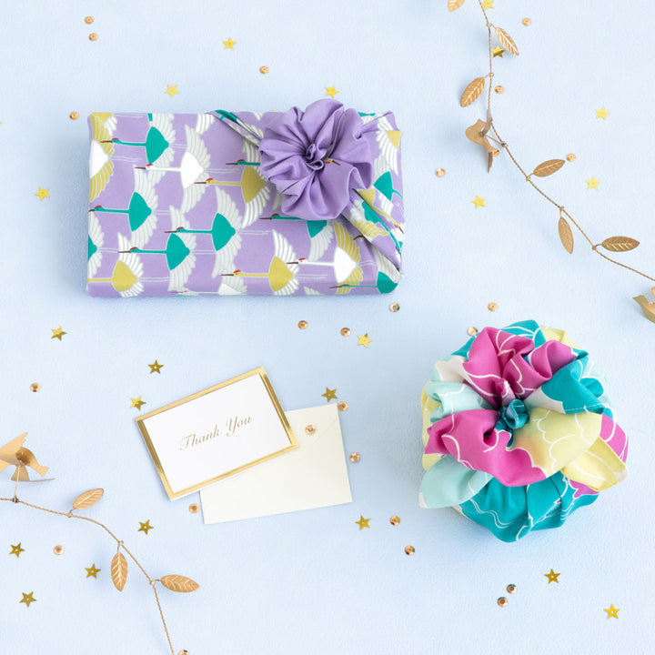 photo d'ambiance d'un emballage cadeau réalisée avec le furoshiki au motif d'envolée de grues dans les tons violet lilas, jaune moutarde et vert menthe adeline x musubi en 50cm par 50cm