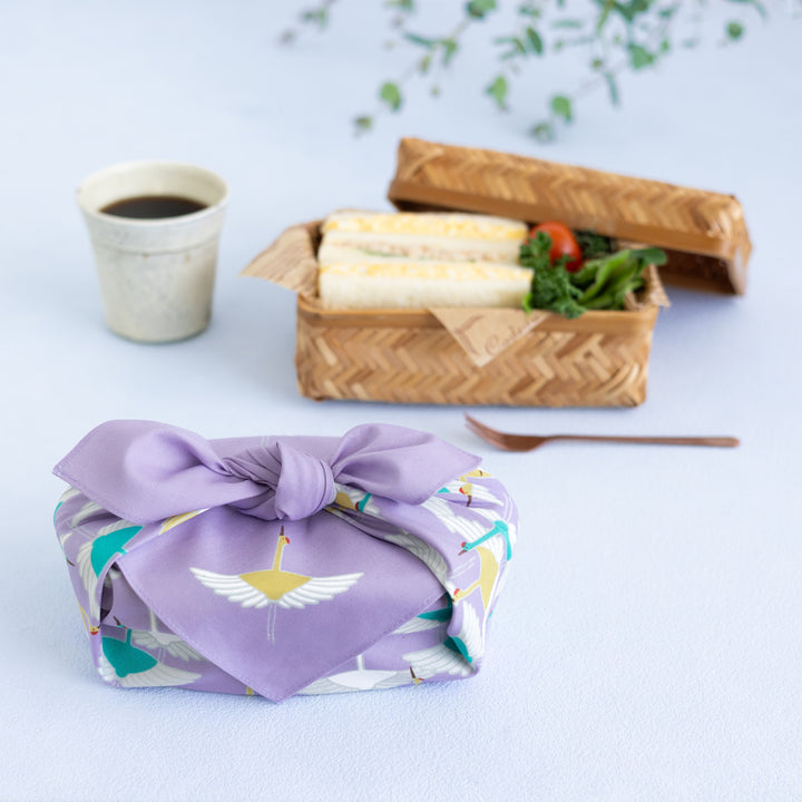 photo d'ambiance d'un emballage cadeau réalisé avec le furoshiki au motif d'envolée de grues dans les tons violet lilas, jaune moutarde et vert menthe adeline x musubi en 50cm par 50cm