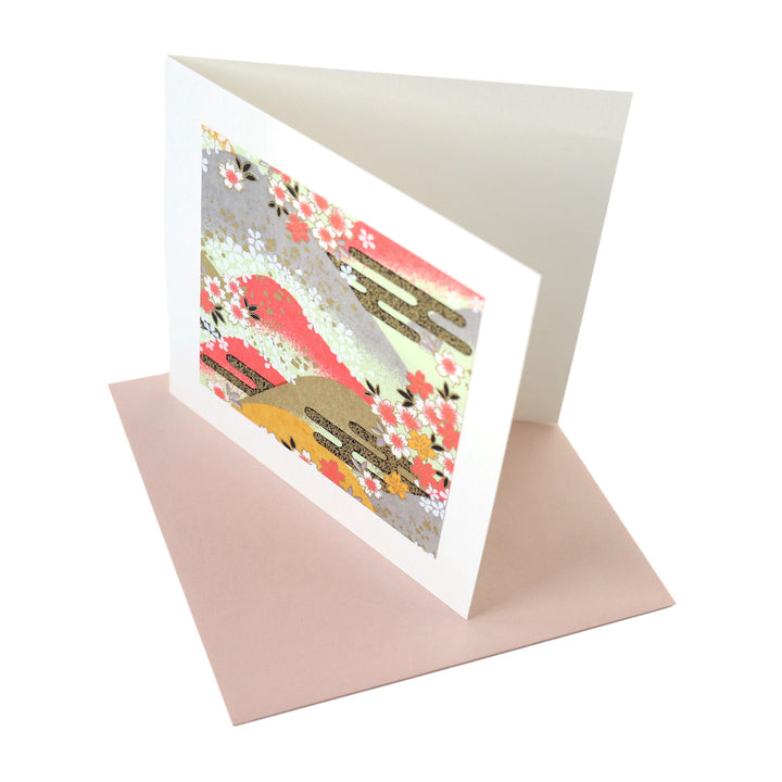 photo packshot vue de biais de la carte carrée double habillée d'un carré de papier japonais de 13cm au motifs de montagnes et de fleurs de cerisier dans les tons roses, jaunes, verts, gris et doré (M1033) et son enveloppe assortie adeline klam