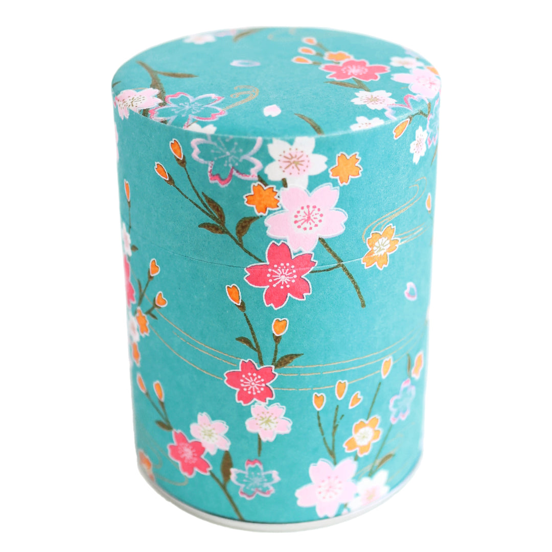 photo packshot d'une boîte à thé cylindrique fermée tapissée d'un papier japonais aux motifs de branches de fleurs de cerisier et d'ondulations dans les tons bleu turquoise, roses et dorés (M380) adeline klam