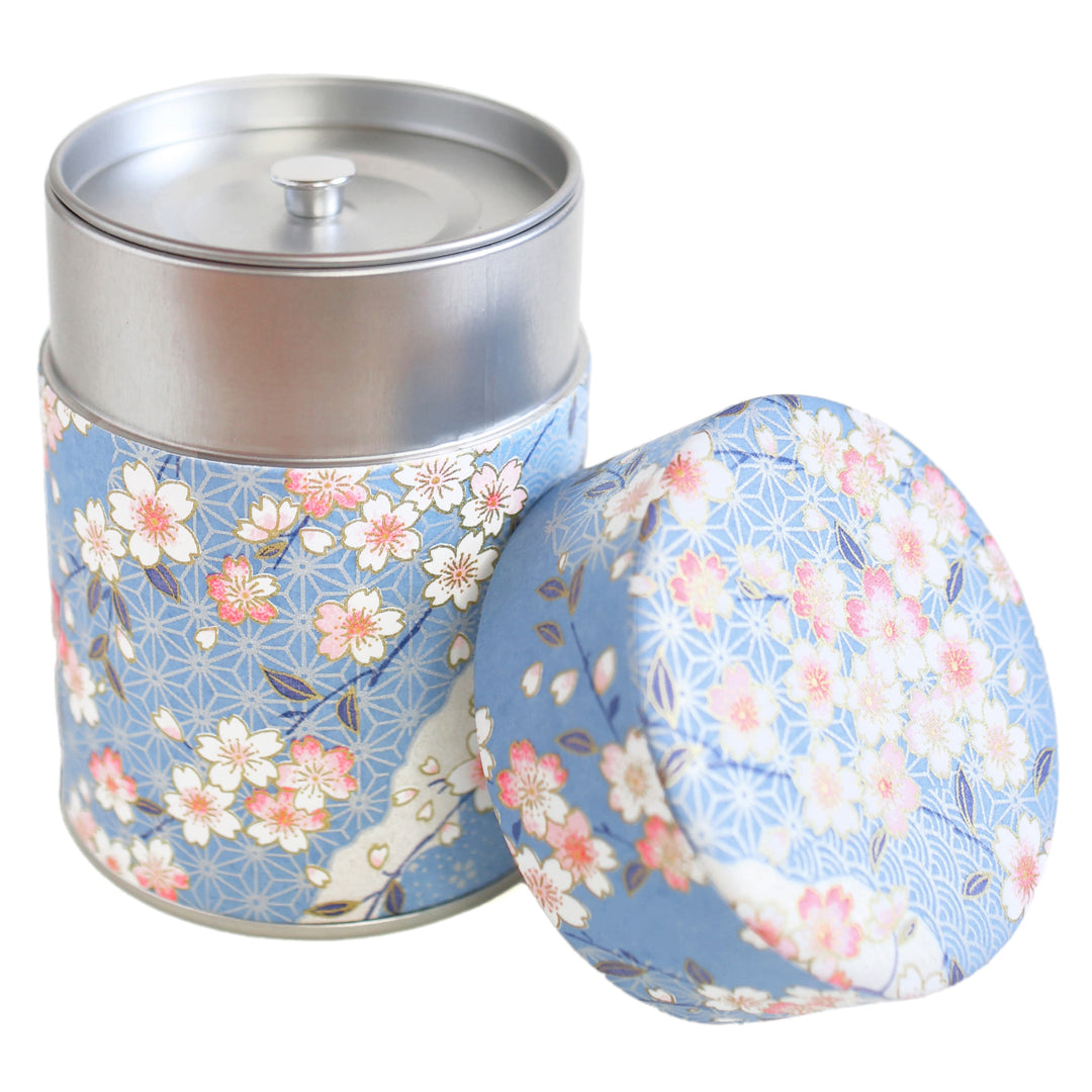 photo packshot d'une boîte à thé cylindrique ouverte tapissée d'un papier japonais aux motifs de fleurs de cerisier, et de petits motifs japonais variés dans les tons bleu clair, roses et blancs (M384) adeline klam
