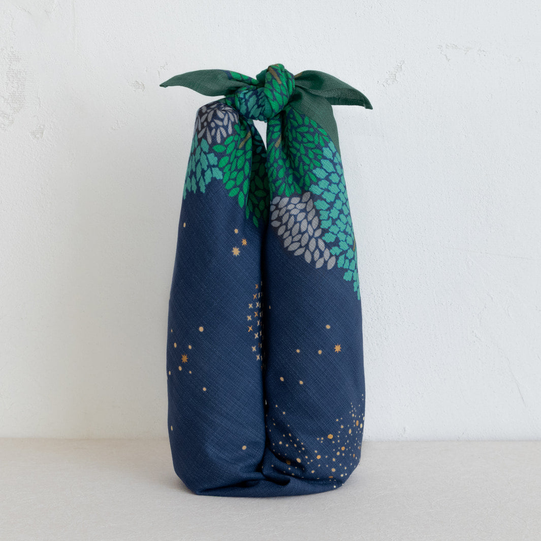 photo d'ambiance de bouteilles de vin emballées avec le furoshiki au motif de forêt étoilée dans les tons bleu marine et turquoise, verts et violet lavande adeline x musubi en 70cm par 70cm