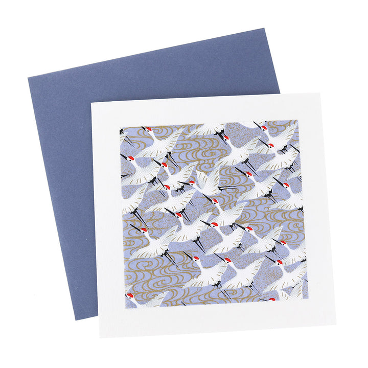 photo packshot vue de face de la carte carrée double habillée d'un carré de papier japonais de 13cm au motifs d'envolée de grues blanches sur fond bleu lavande (M388) et son enveloppe bleu foncé adeline klam