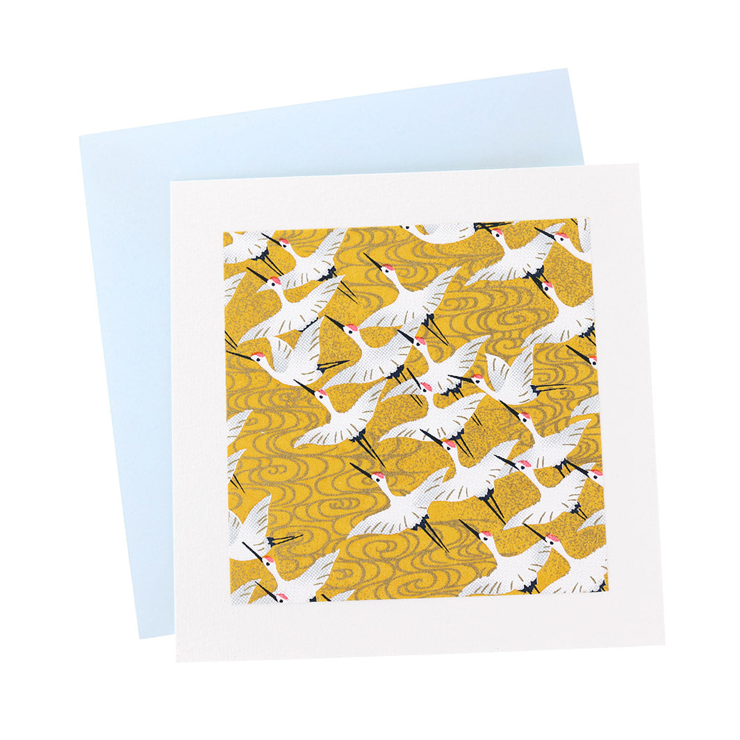 photo packshot vue de face de la carte carrée double habillée d'un carré de papier japonais de 13cm au motifs d'envolée de grues blanches sur fond jaune moutarde (M930) et son enveloppe bleu ciel adeline klam