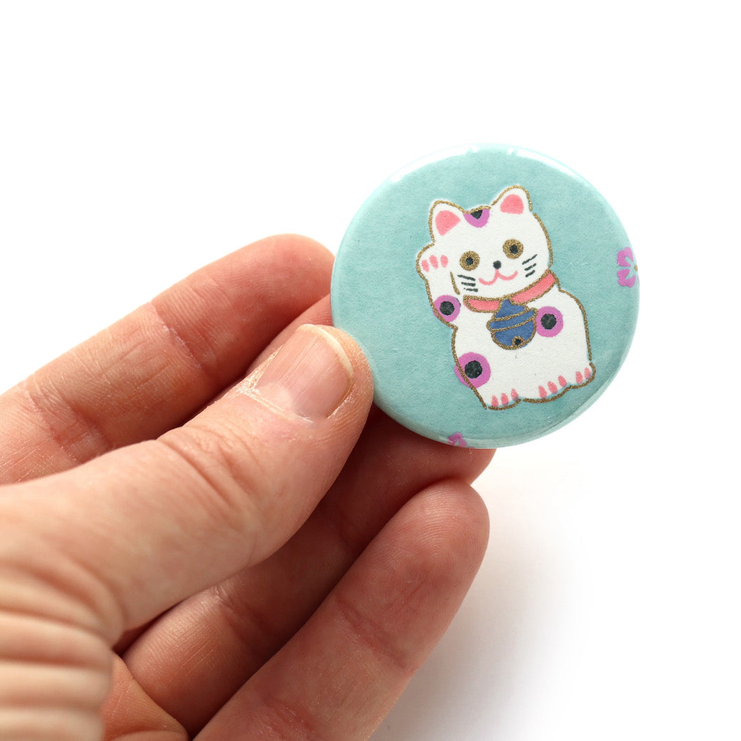 photo packshot en main du recto de l'aimant en papier japonais de 45mm de diamètre aux motifs de maneki-neko blancs aux détails mauve et corail sur fond vert d'eau (M837) adeline klam