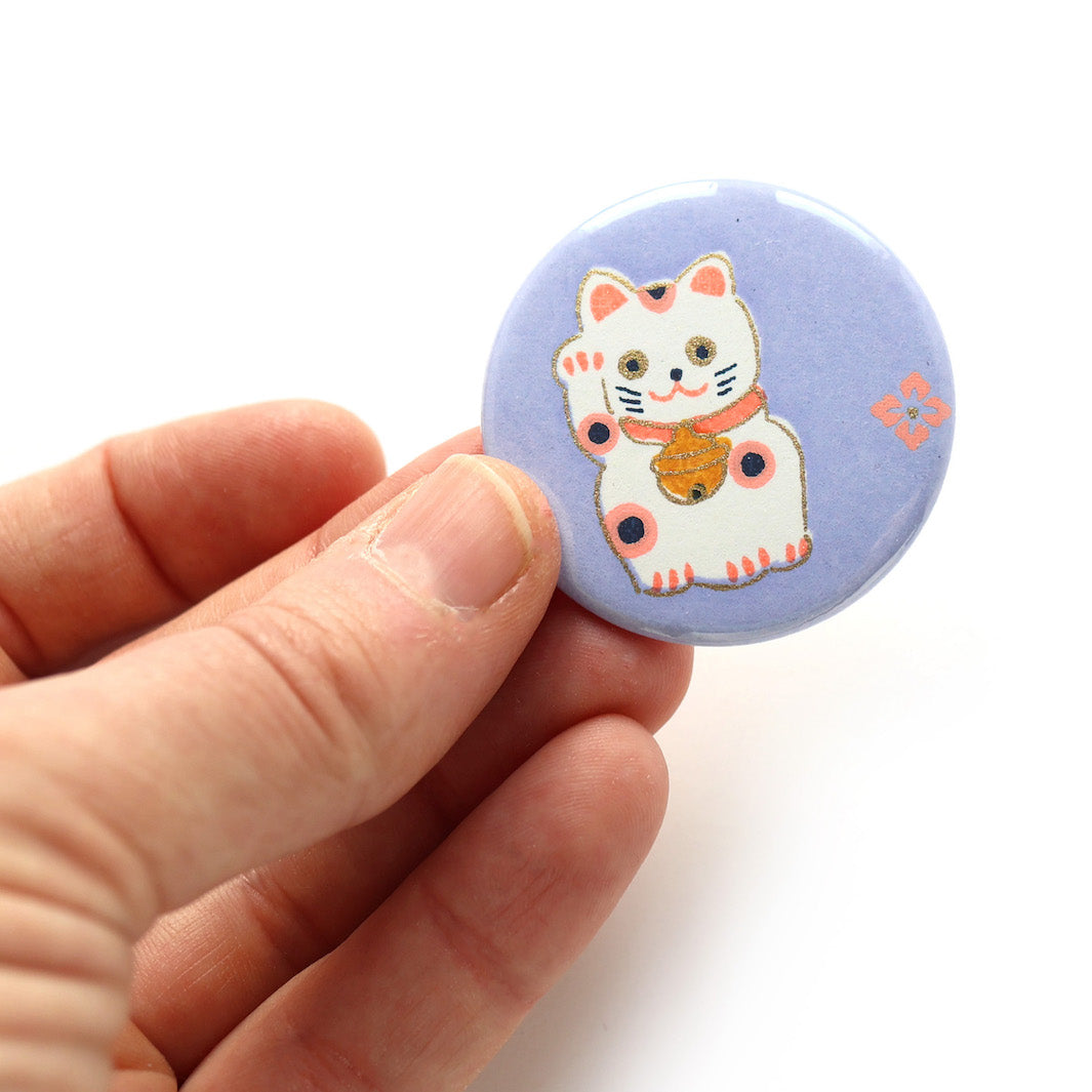 photo packshot en main du recto de l'aimant en papier japonais de 45mm de diamètre aux motifs de maneki-neko blancs aux détails roses, corail et jaune moutarde sur fond violet lilas (M839) adeline klam
