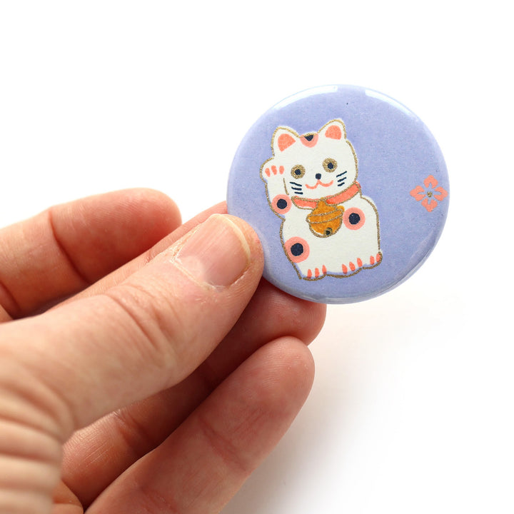 photo packshot en main du recto de l'aimant en papier japonais de 45mm de diamètre aux motifs de maneki-neko blancs aux détails roses, corail et jaune moutarde sur fond violet lilas (M839) adeline klam